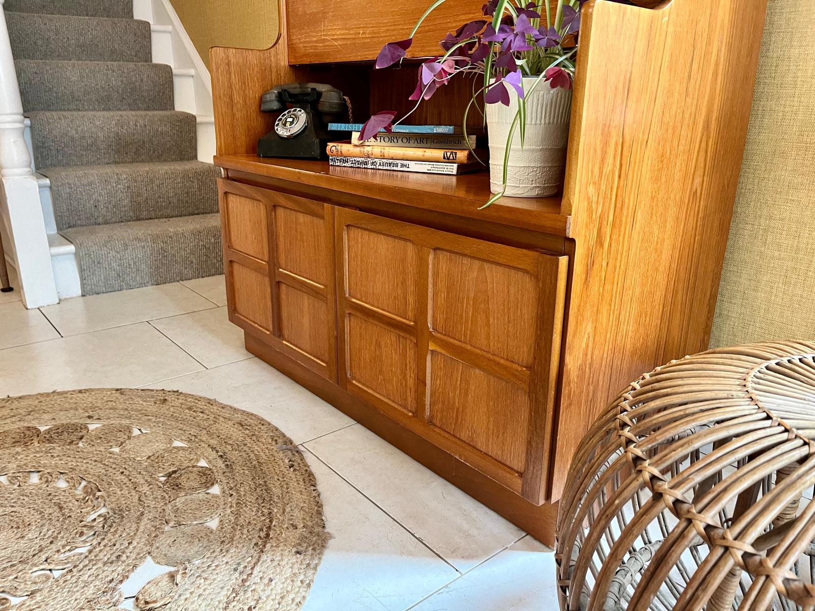 A Nathan mid century drinks cabinet with a drop-down wooden countertop, shown in a home setting next to a staircase. The cabinet is styled with liquor bottles, crystal glasses, and a small shelf inside by Grace & Joy Furniture