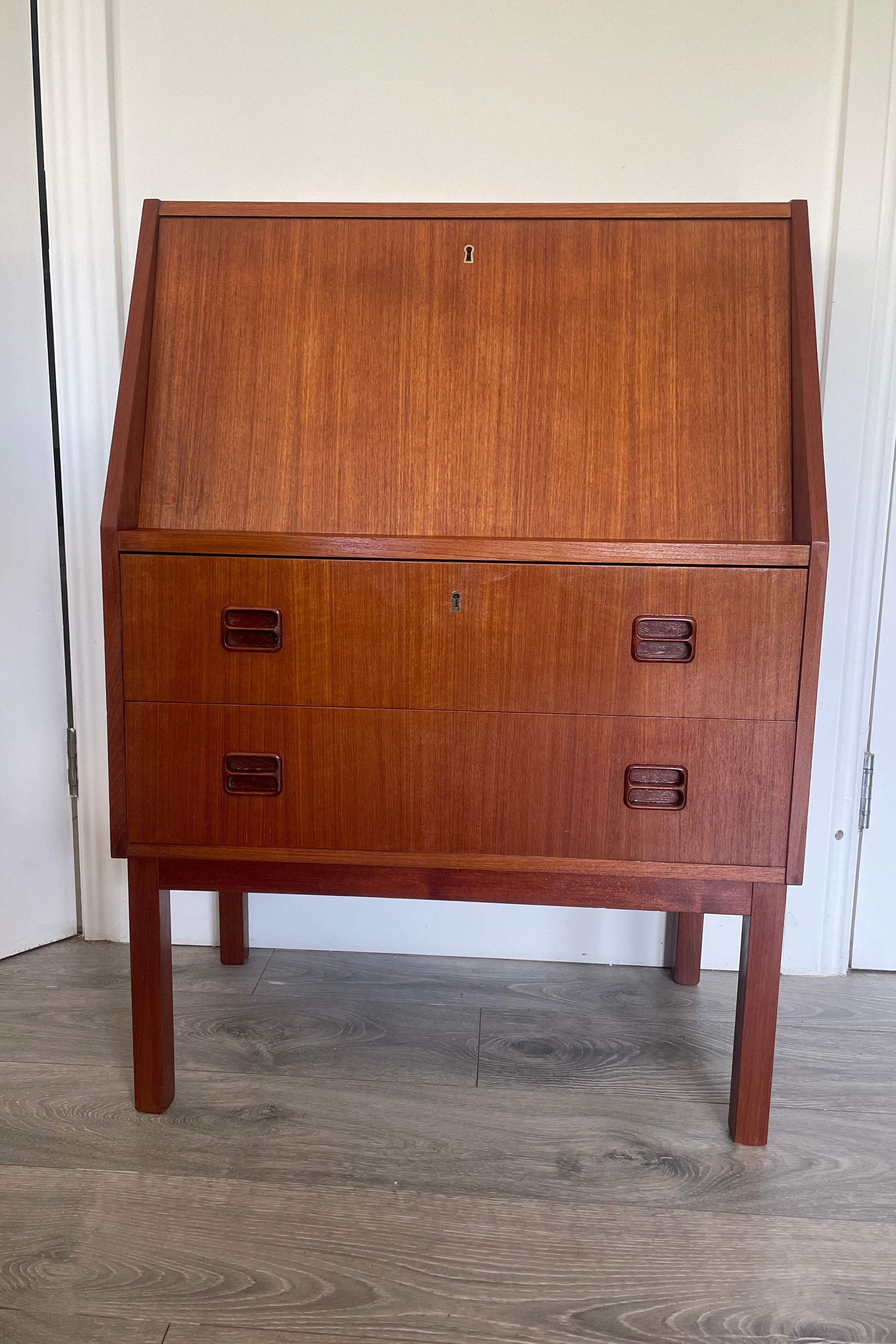 A mid-century modern Danish bureau with a warm reddish-brown finish, featuring a large left door and three drawers on the right with dark metal pulls, photographed indoors on a gray wood floor. by Grace & Joy Furniture