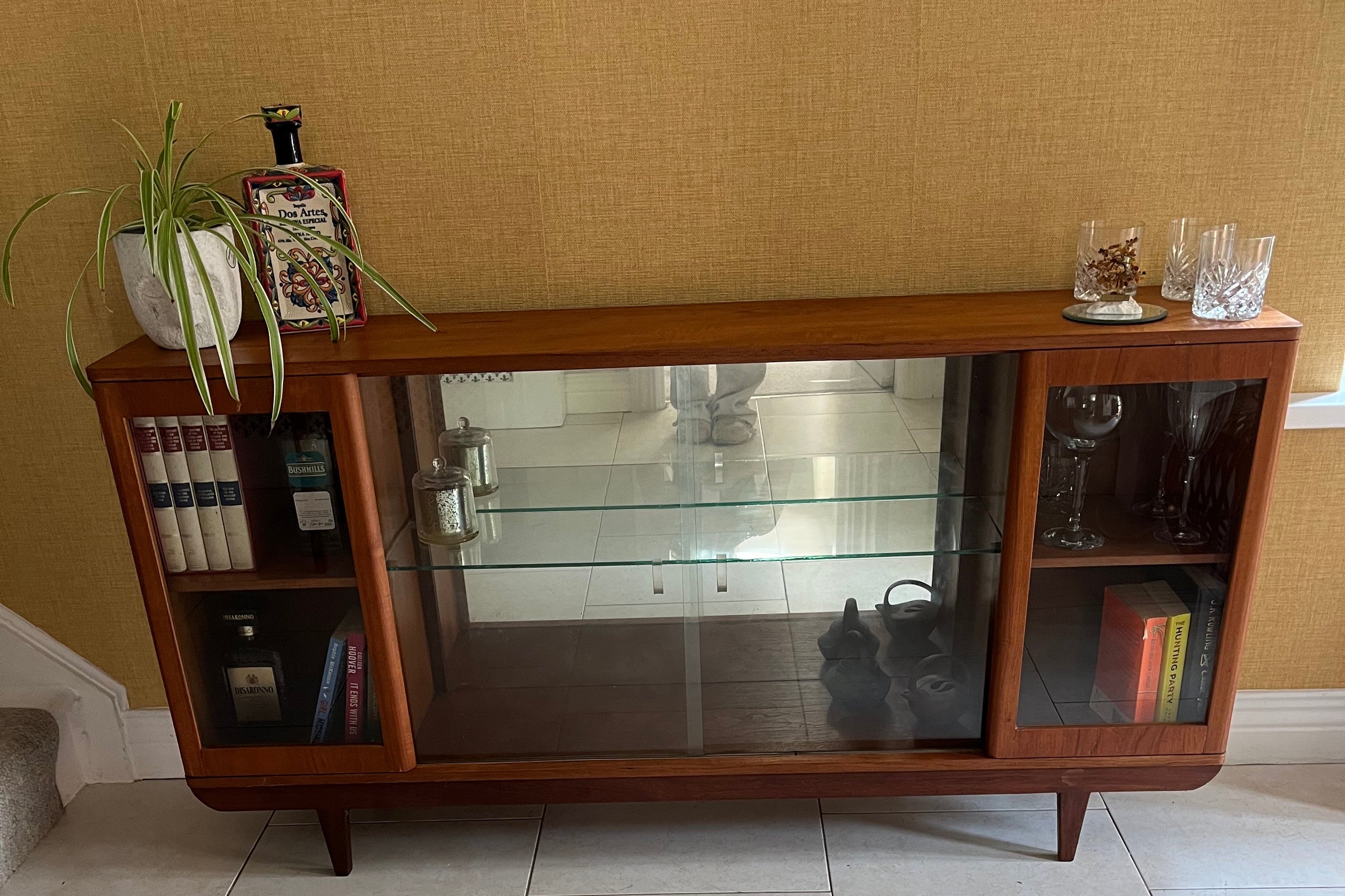A mid-century modern SF Quinn wooden display cabinet with glass doors, shown in a home interior setting with tiled floors and textured walls by Grace & Joy Furniture