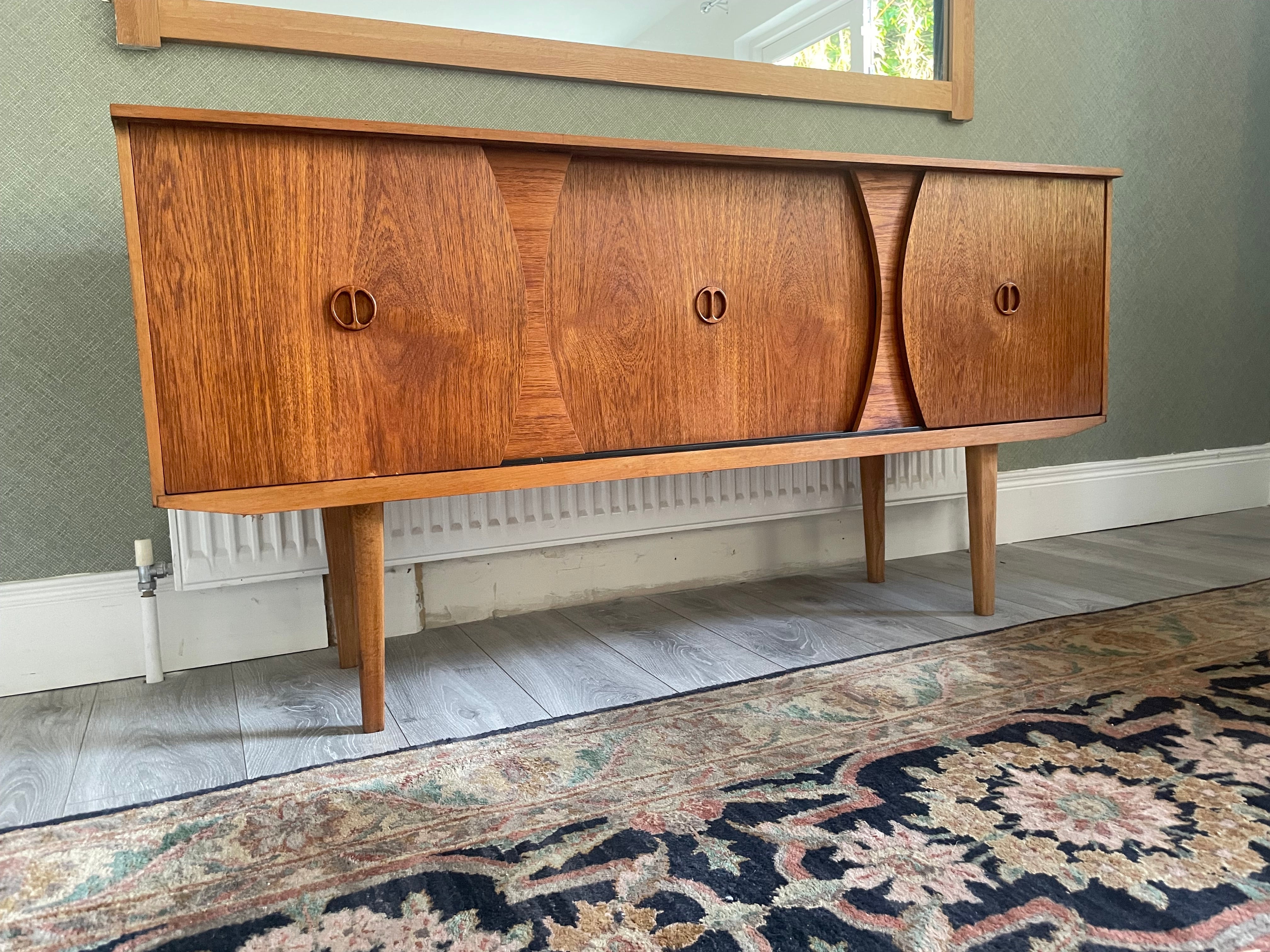Restored mid-century modern wooden credenza or cabinet with three curved doors and circular pull handles, photographed in a home interior with wood flooring and a patterned rug visible by Grace & Joy Furniture