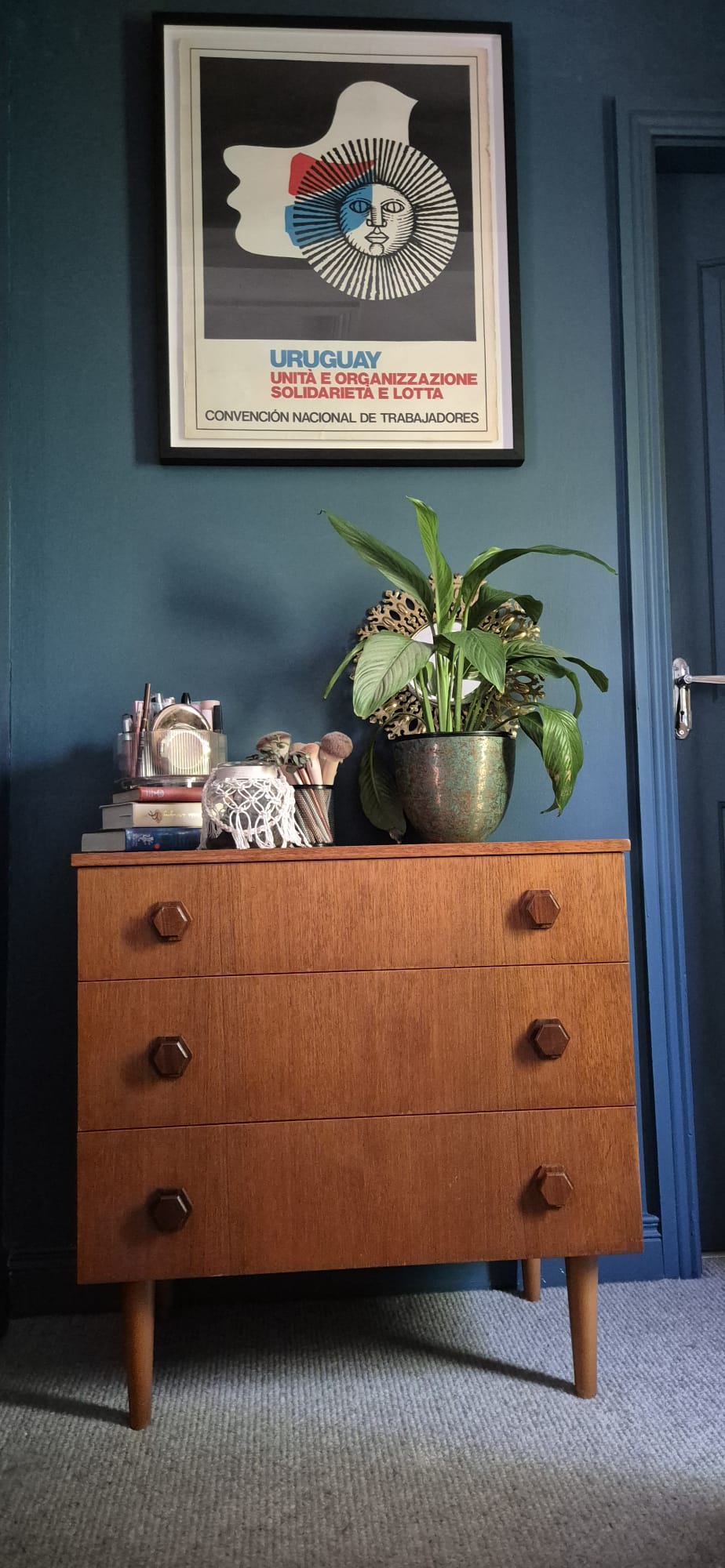 A mid-century modern wooden chest of drawers with three drawers and round dark knobs, styled with a potted plant, books, and a small decorative figure in a room with a dark blue wall by Grace & Joy Furniture