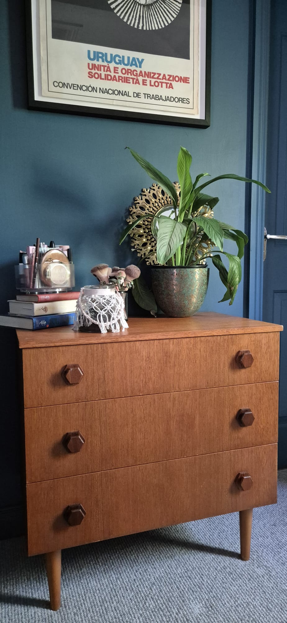 A mid-century modern wooden chest of drawers with three drawers and round dark knobs, styled with a potted plant, books, and a small decorative figure in a room with a dark blue wall by Grace & Joy Furniture