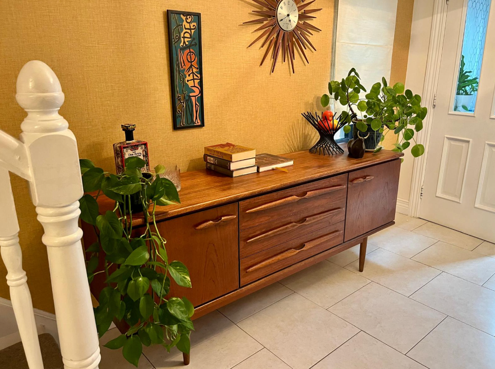 A mid-century modern wooden sideboard with a warm medium brown finish, styled in a home entryway with potted green plants, books and a sunburst wall clock.