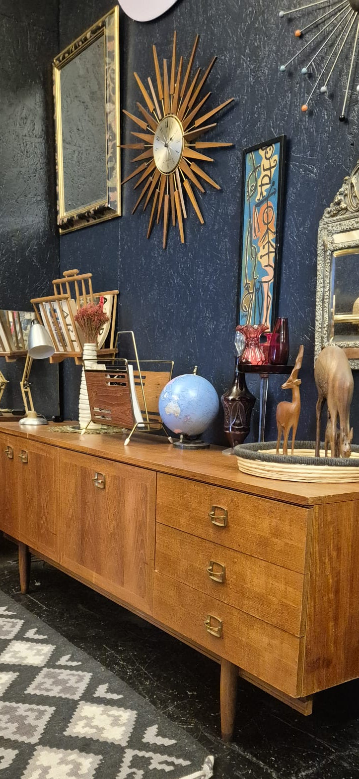A mid-century modern wooden sideboard cabinet with six drawers, styled in a dark room with a patterned rug, decorative mirrors, and wall art.