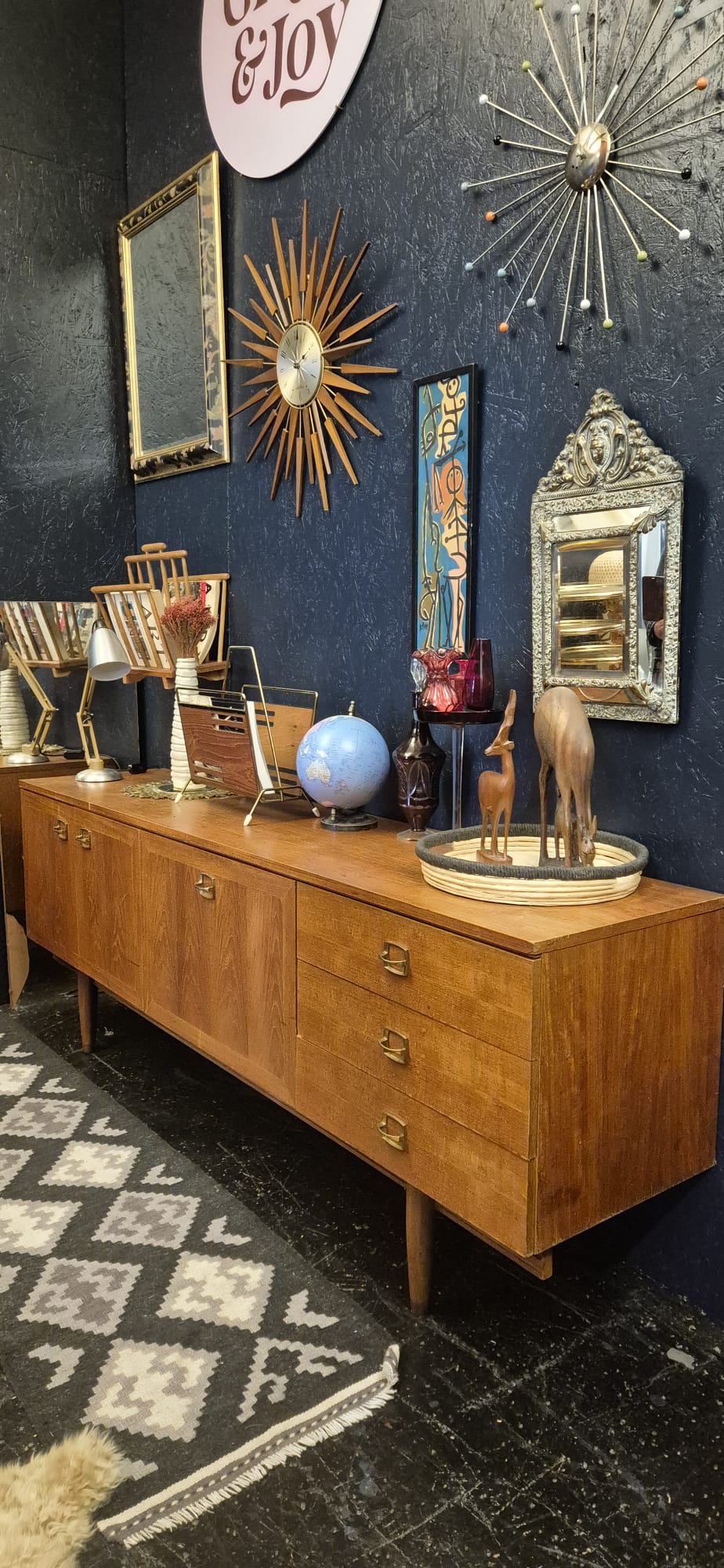 A mid-century modern wooden sideboard console cabinet with six drawers and tapered legs, styled in a bohemian home decor showroom against a dark blue wall.