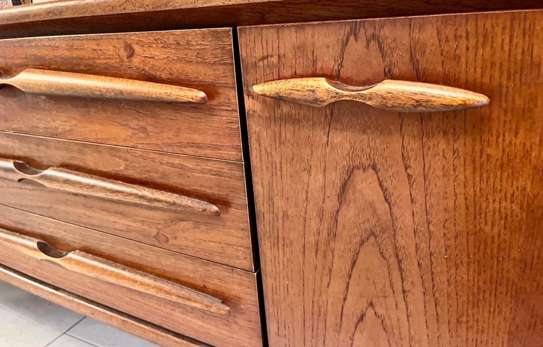 A mid-century modern wooden sideboard with a warm medium brown finish, styled in a home entryway with potted green plants, books and a sunburst wall clock.