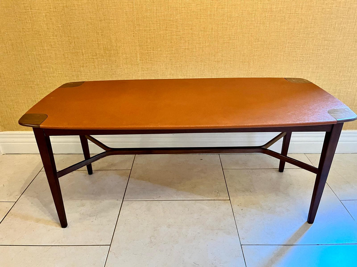 A mid-century modern coffee table with a warm brown leather top and dark wood legs, set against a textured beige wall and light tile floor.