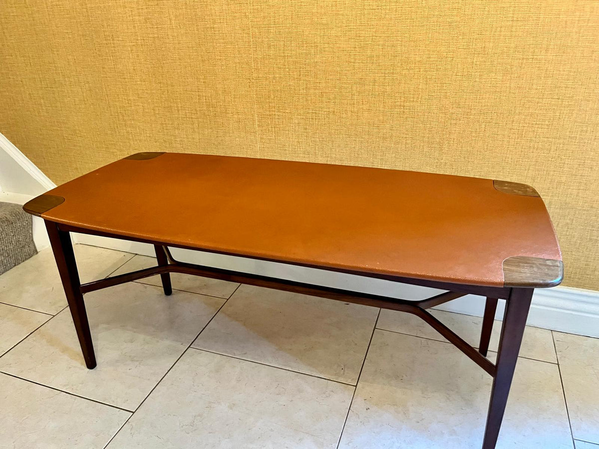 A mid-century modern wooden coffee table with a warm orange-brown leather top and dark wood legs, set against a textured beige wall and tiled floor.