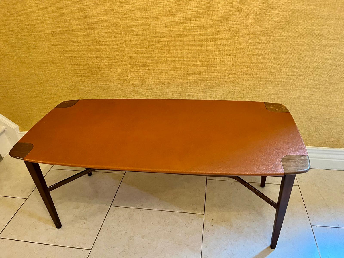 A mid-century modern rectangular coffee table with a warm orange-brown wood top, dark brown angled legs, and metal corner protectors, set against a textured yellow wall and tiled floor.