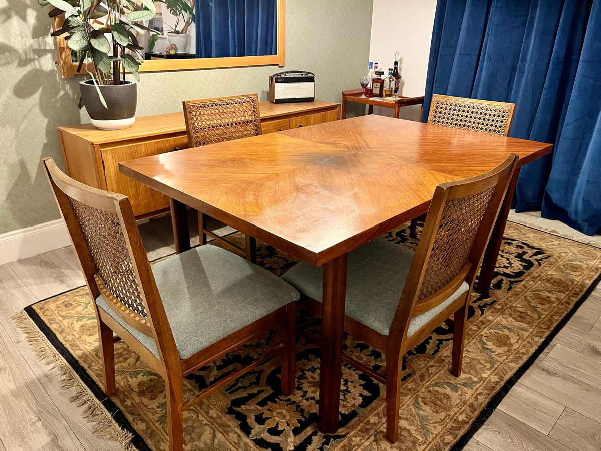 A mid-century modern wooden dining table with four matching woven-back chairs, set on a patterned area rug in a dining room.