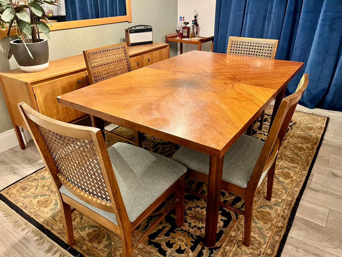 A mid-century modern wooden dining table set with six matching chairs featuring woven cane backs and light gray upholstered seats, placed on a patterned area rug in a dining room.