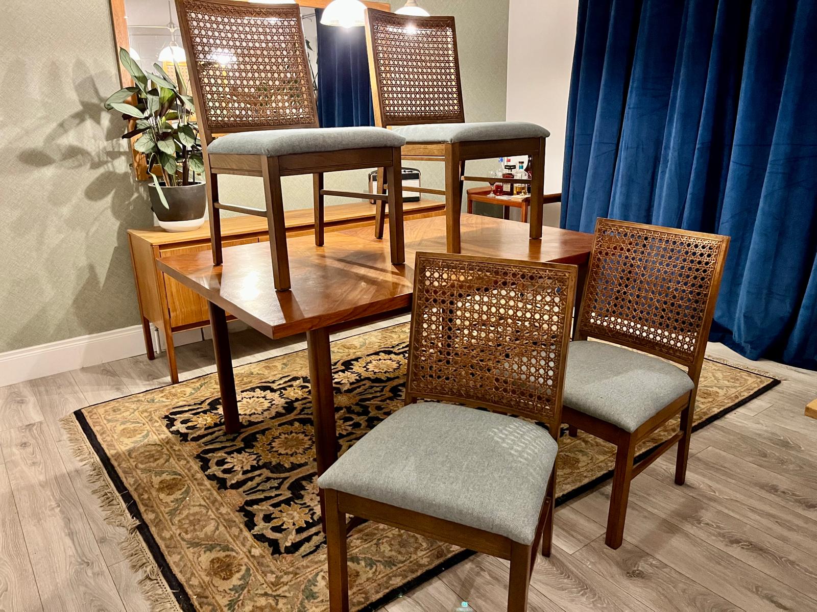 A wooden dining table with four matching chairs featuring woven cane backs and light gray upholstered seats, arranged on a patterned area rug in a furnished room with blue curtains and a mirror.
