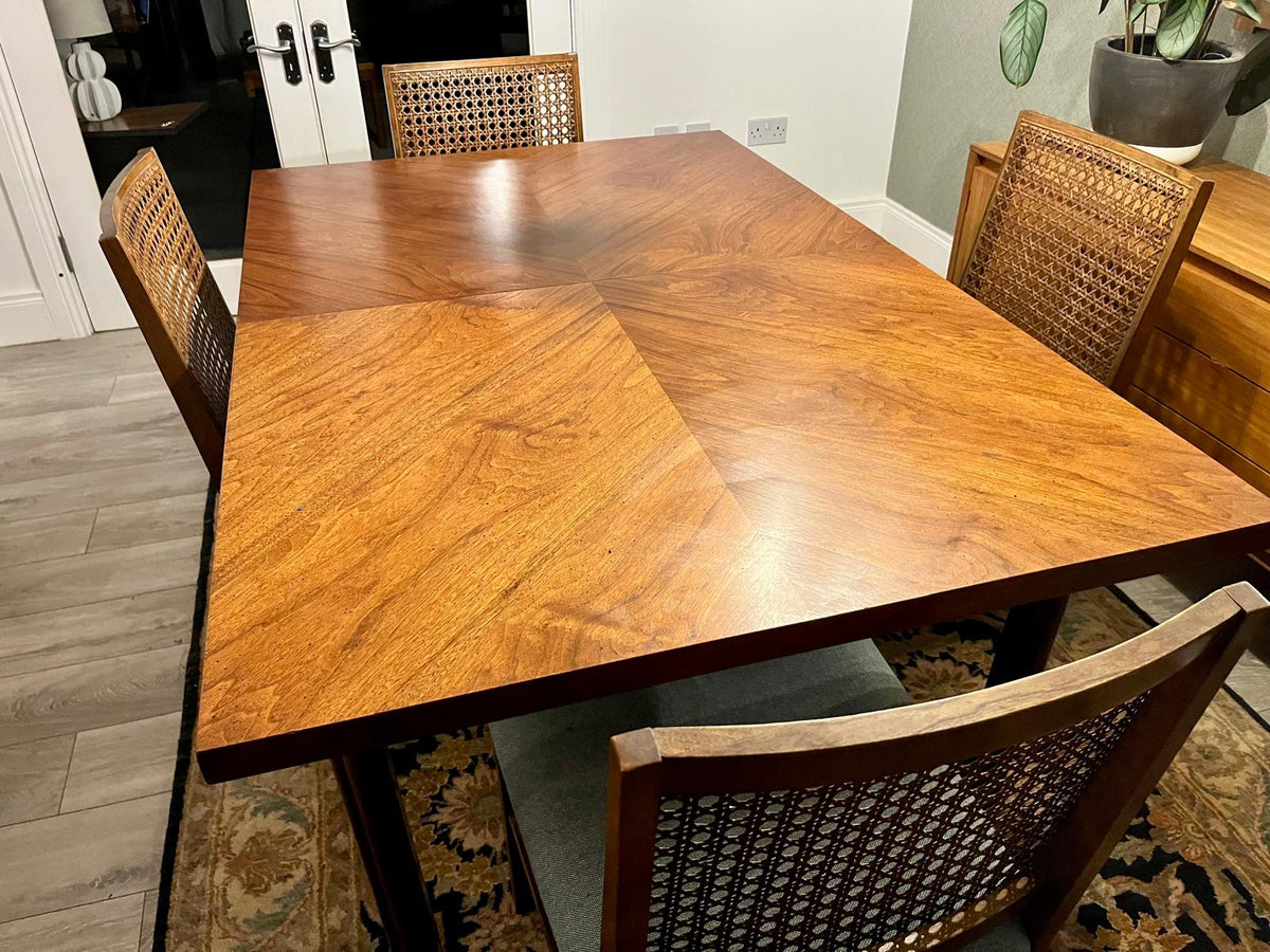 A rectangular wooden dining table with a warm medium-brown finish and matching woven-back chairs, shown in a real indoor room with wood flooring, a rug, and wall decor.