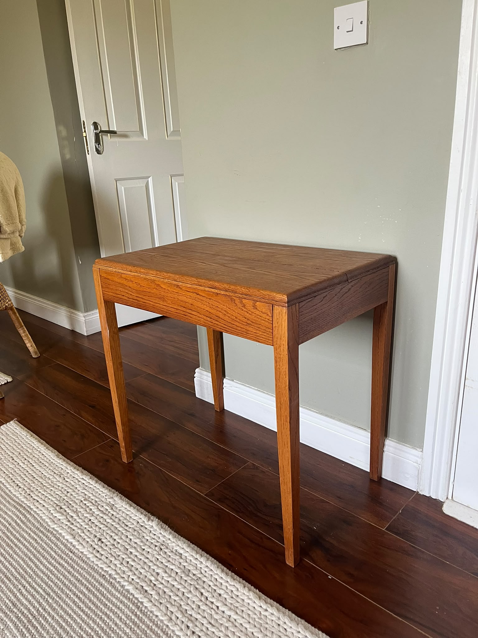 A mid-century modern wooden side table with a warm medium-brown finish, styled with a green pleated table lamp and a stack of books in a room with dark wood floors and a sage green wall by Grace & Joy furniture