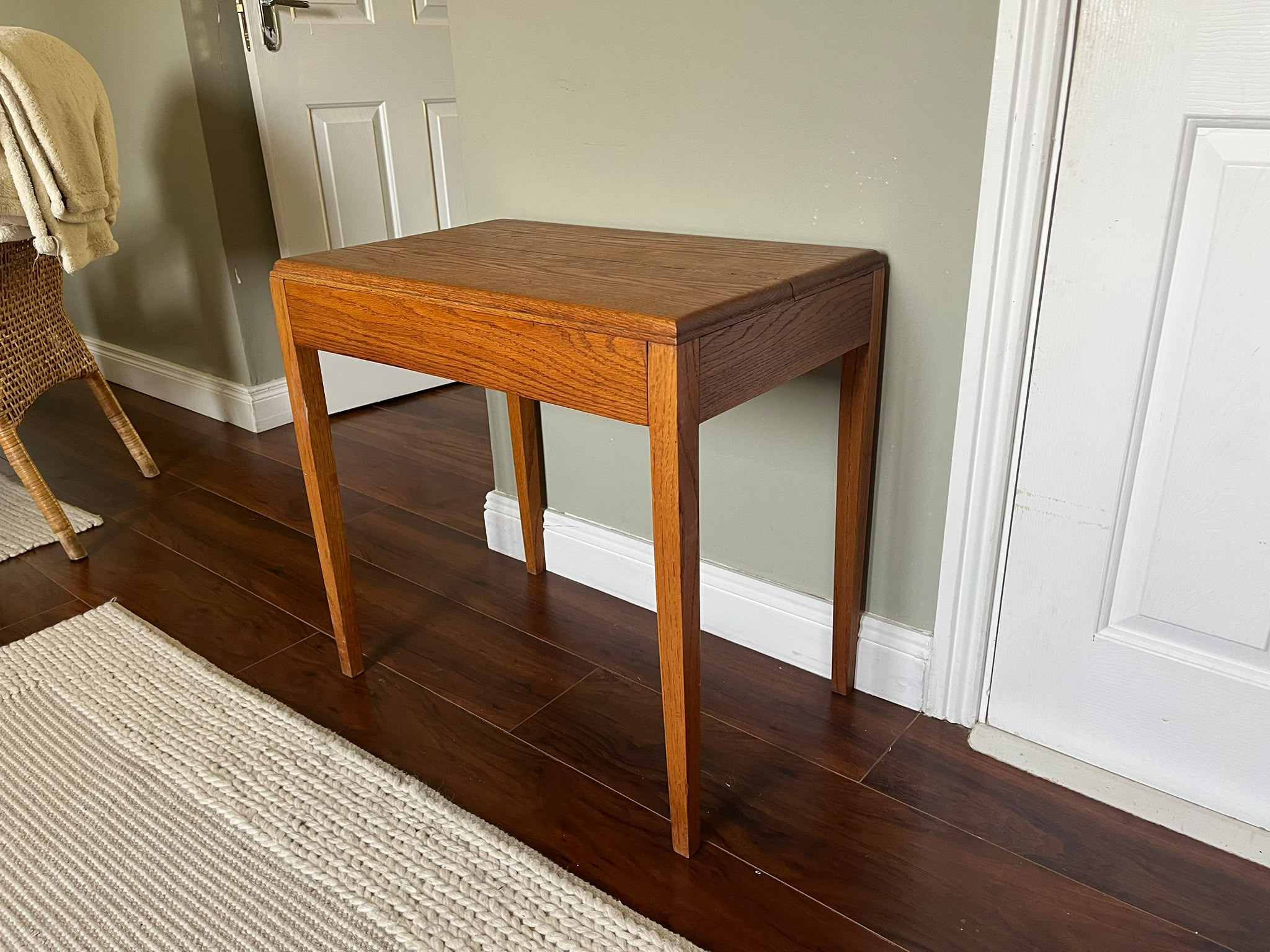 A mid-century modern wooden side table with a warm medium-brown finish, styled with a green pleated table lamp and a stack of books in a room with dark wood floors and a sage green wall by Grace & Joy furniture