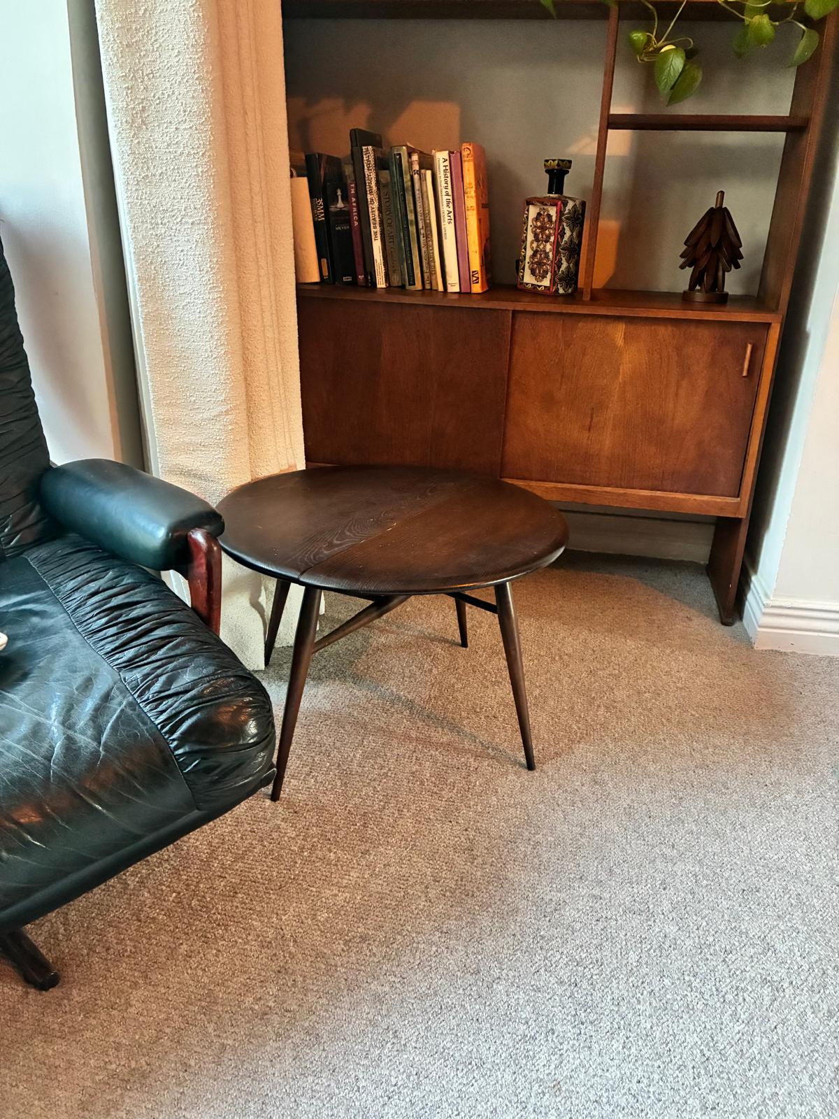 A mid-century modern ercol wooden side table with a round dark top and angled legs, positioned in a room with a beige carpet, a curtain, and a mid-century credenza in the background.