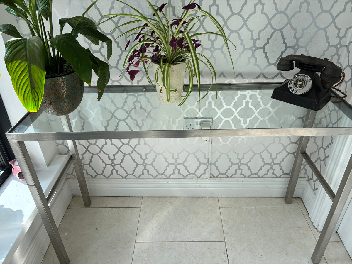 A modern console table with a clear glass top and brushed silver metal frame, styled with potted plants and a vintage black rotary telephone against a white patterned wallpaper background.