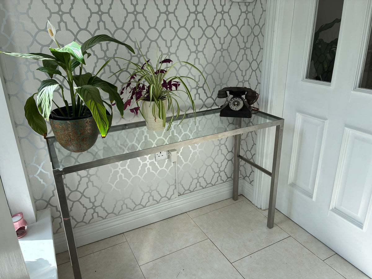 A modern glass-top console table with a silver metal frame, styled with two potted plants and a black rotary telephone, set against a white paneled door and patterned wallpaper.