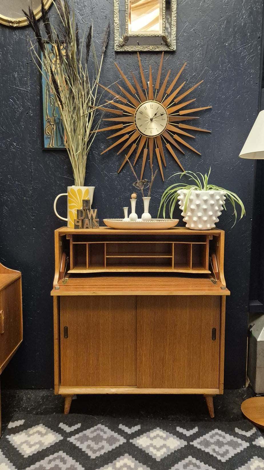 A mid-century modern wooden writing desk or cabinet with a roll-top design, styled in a room against a dark textured wall with decorative mirrors, a sunburst clock, and plants.