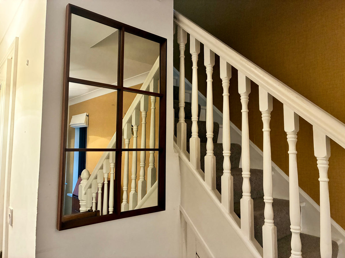 A large rectangular teak wall mirror with a dark wood frame hangs on a white wall beside a staircase with white balusters and a mustard-yellow wall.