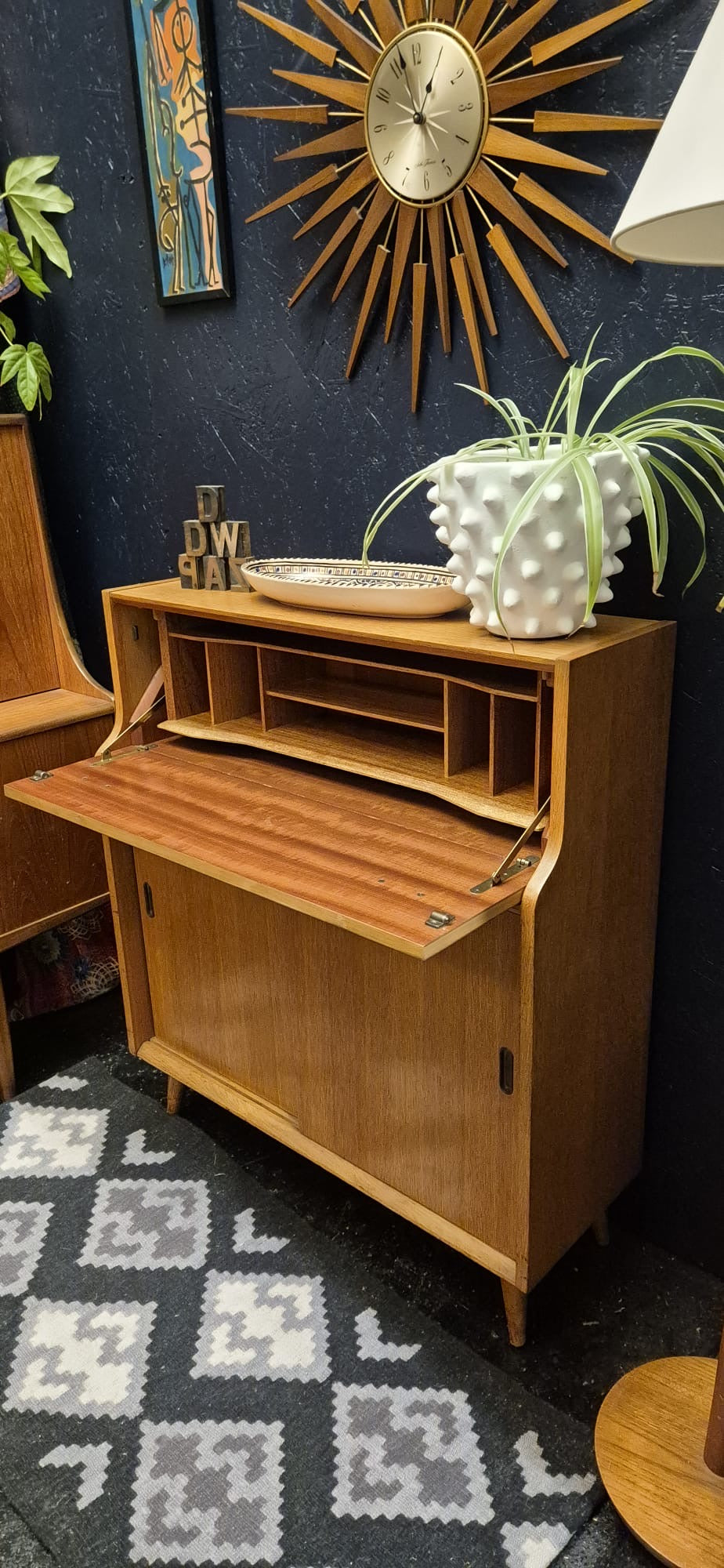A mid-century modern wooden roll-top secretary cabinet with its front door open, styled in a room with a dark blue wall, a sunburst wall clock, a patterned rug, and a potted plant on top.