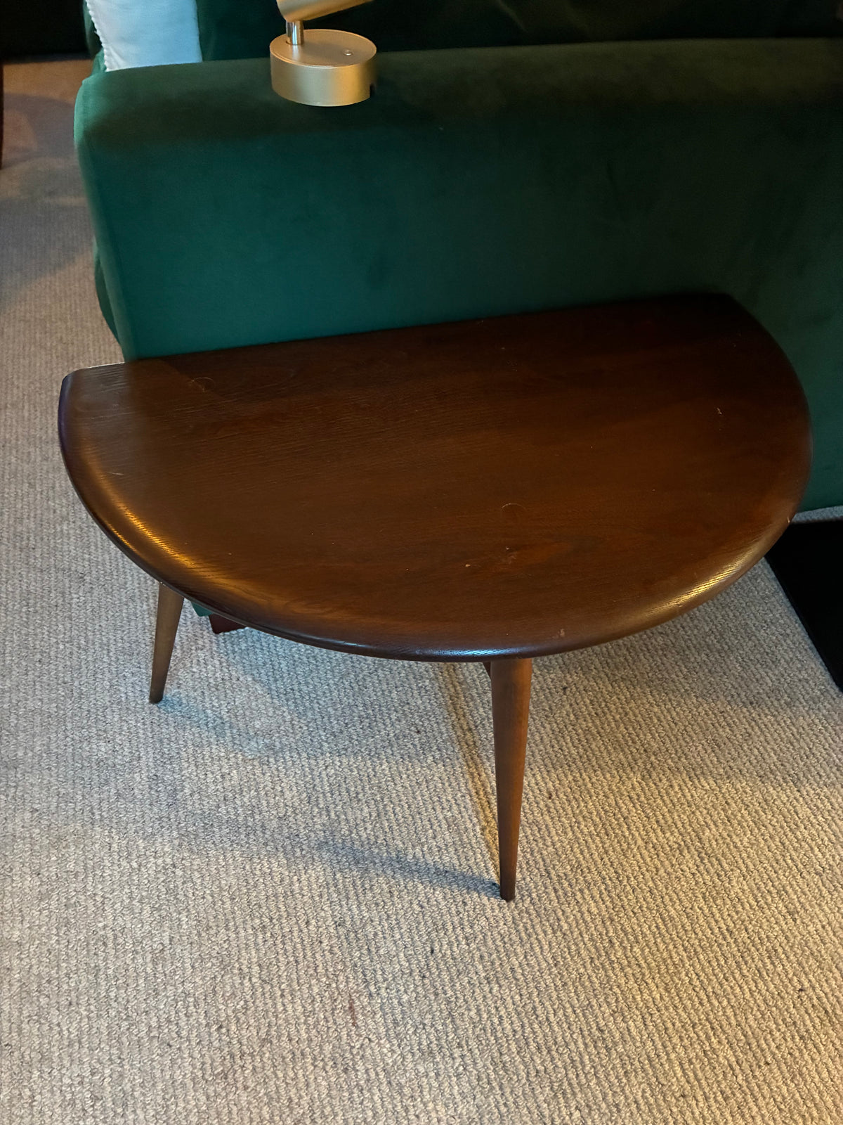 A mid-century modern ercol wooden side table with a semi-circular dark brown top and three tapered legs, positioned in front of a green velvet sofa on a textured beige carpet.