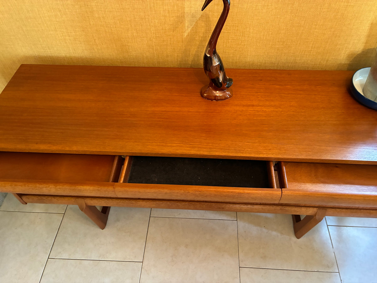 A mid-century modern wooden console table with a warm reddish-brown finish, featuring an open drawer and a decorative metal crane sculpture on top, set against a textured yellow wall and tiled floor.
