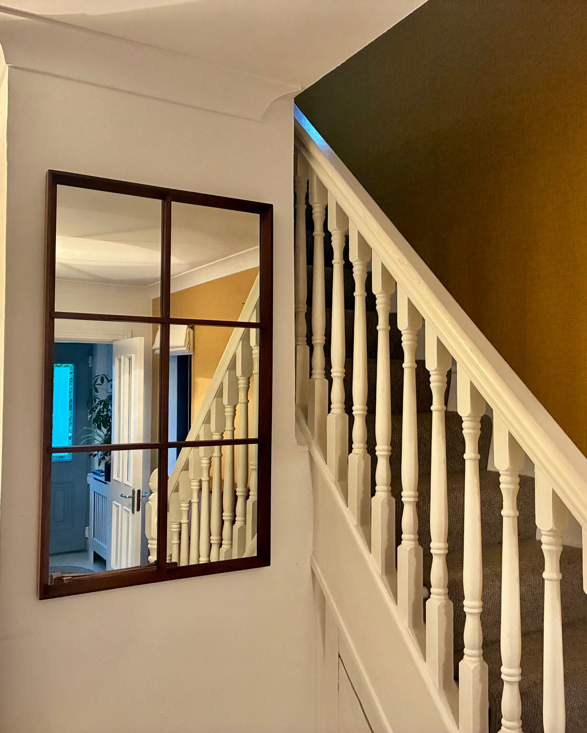 A large rectangular teak wall mirror with a dark wood frame hangs on a white wall beside a staircase with white balusters and a mustard-yellow wall.