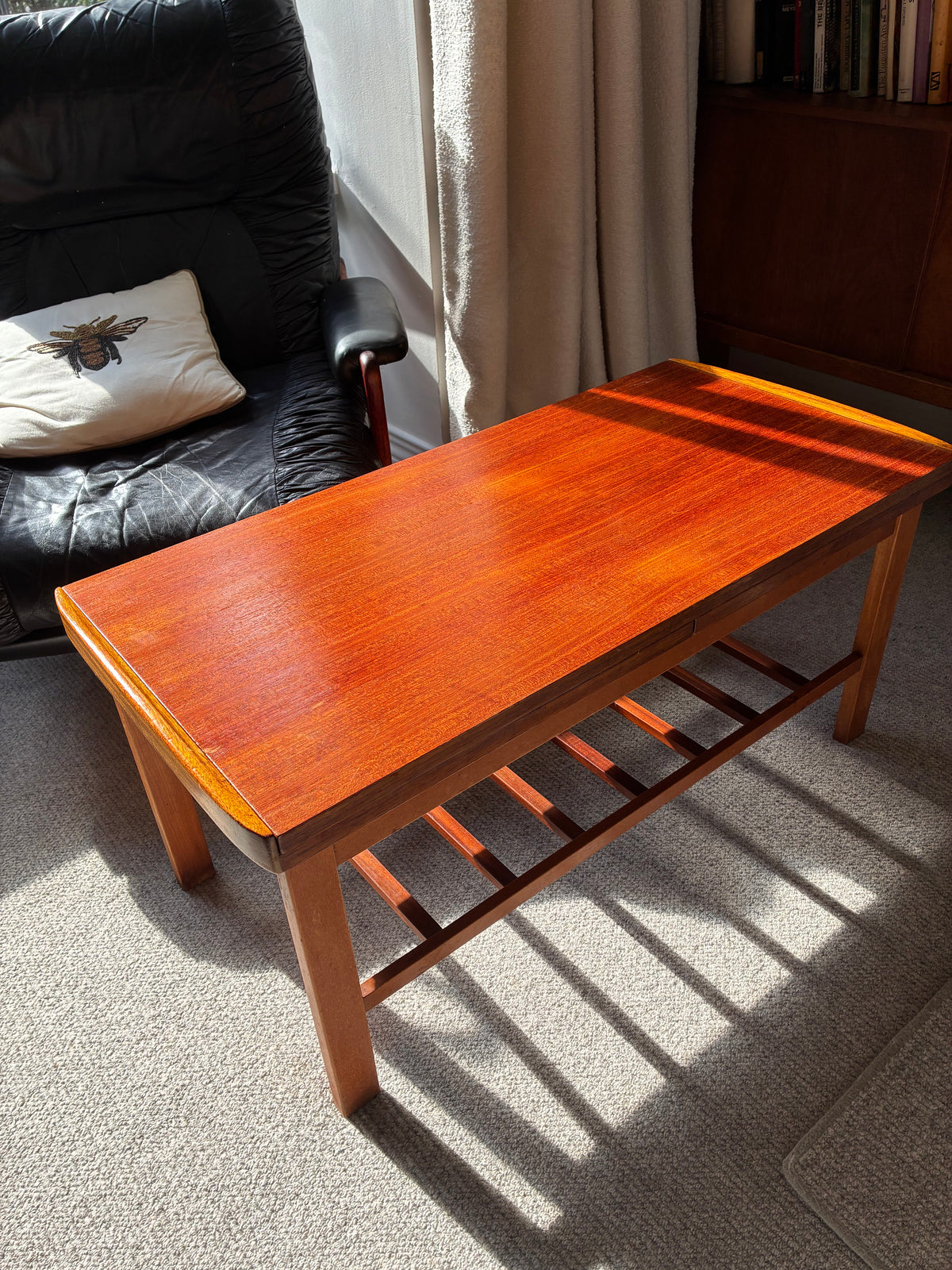A rectangular wooden coffee table with a warm reddish-brown finish and a slatted lower shelf, photographed in a real room with sunlight, a black leather chair, curtains, and carpet visible.
