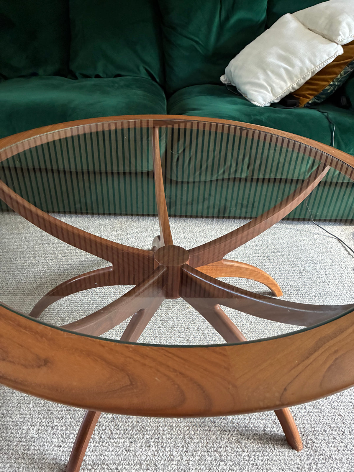 A mid-century modern g plan spider wooden coffee table with a clear glass top and curved star-shaped legs, positioned in a living room in front of a green velvet sofa.