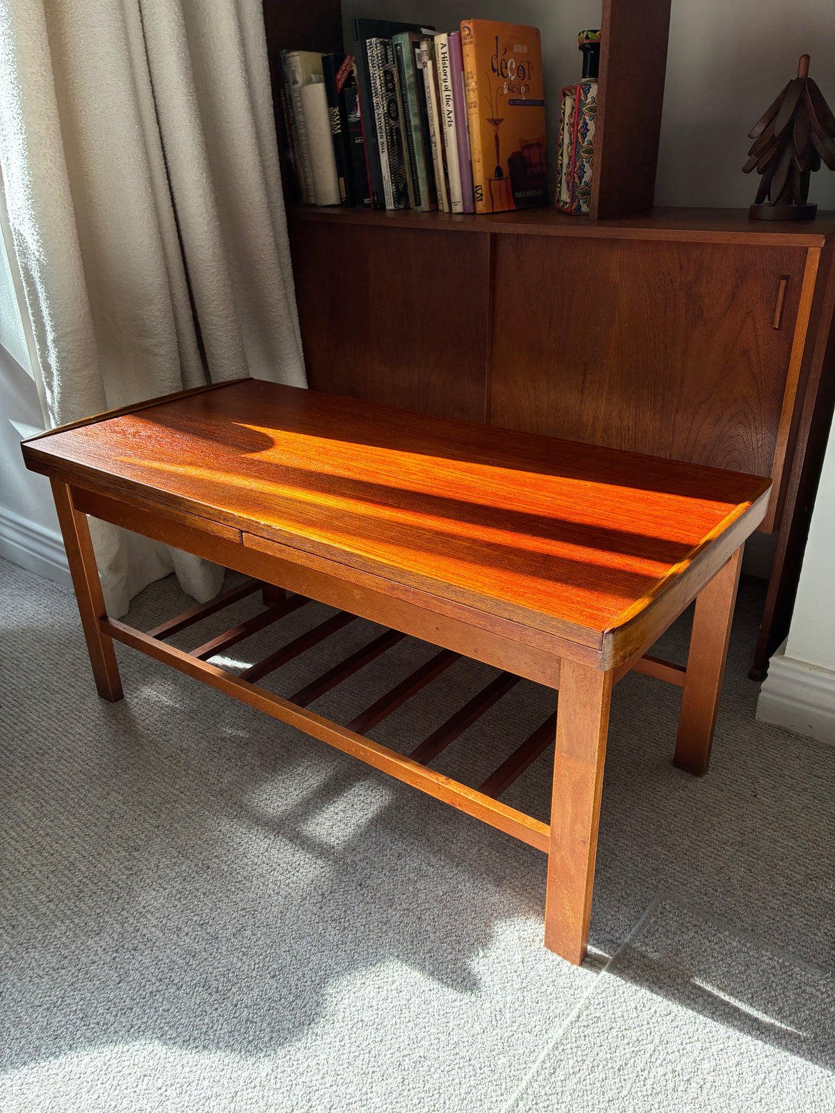 A rectangular wooden coffee table with a glossy reddish-brown finish and a slatted lower shelf, placed in a room with a bookshelf, curtains, and carpet visible.