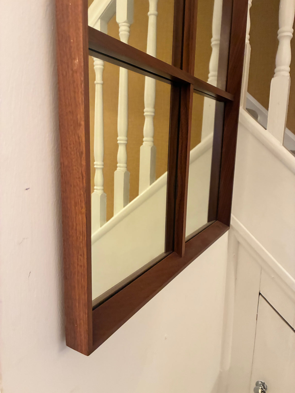 A large rectangular teak wall mirror with a dark wood frame hangs on a white wall beside a staircase with white balusters and a mustard-yellow wall.
