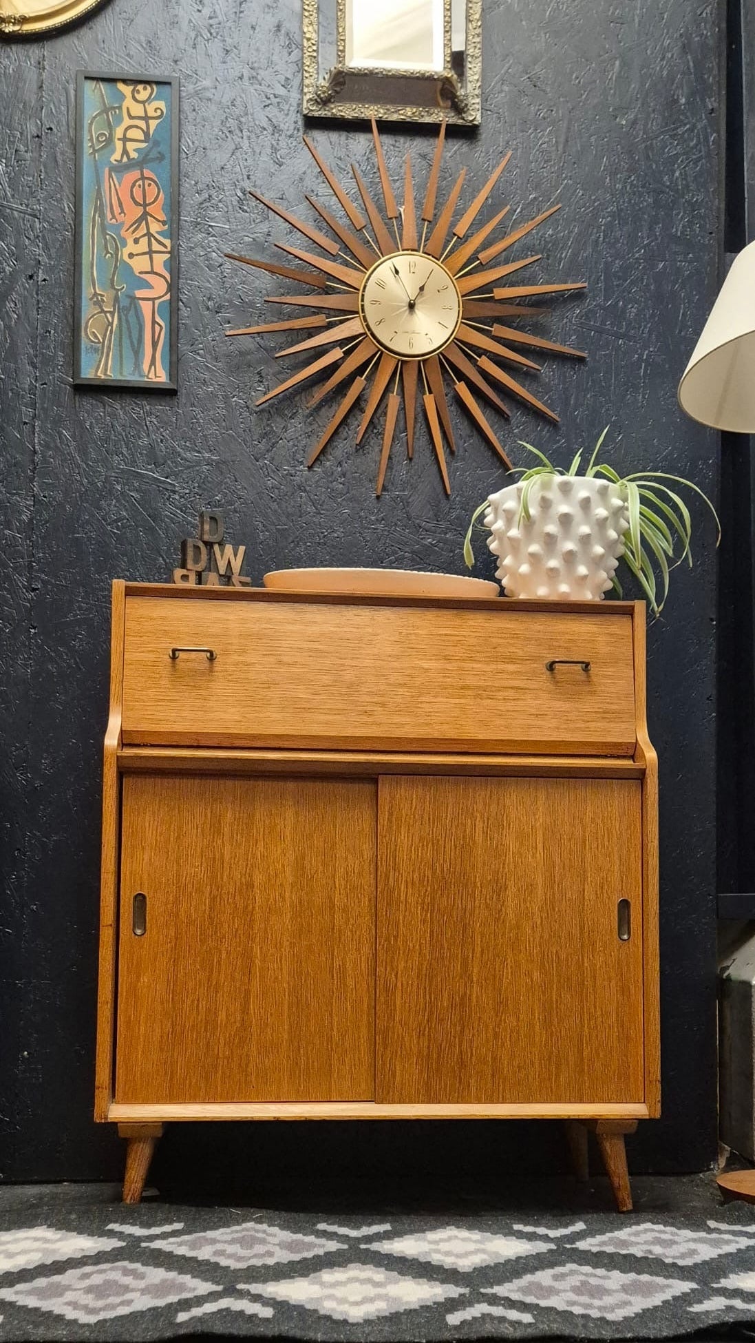 A mid-century modern wooden cabinet with a warm honey finish, styled in a room against a dark textured wall with a sunburst clock, framed art, and a patterned rug.