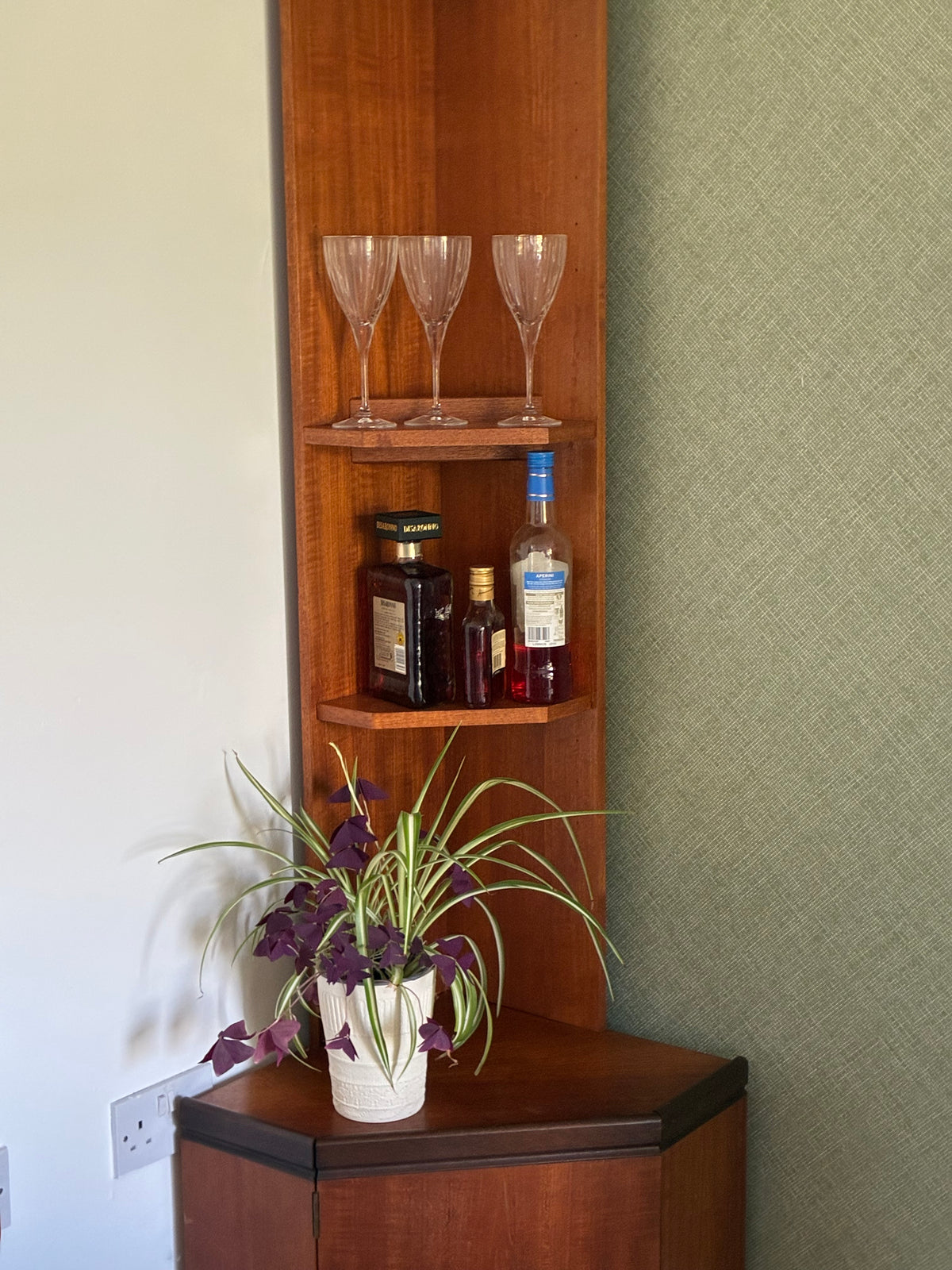 A corner wooden g plan shelf cabinet with two open shelves, holding two wine glasses, two liquor bottles, and a potted spider plant on the lower corner cabinet. The cabinet is set against a textured sage green wall and a plain off-white wall.