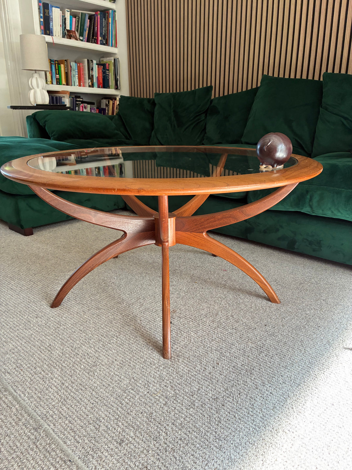 A mid-century modern g plan spider wooden coffee table with a clear glass top and curved star-shaped legs, positioned in a living room in front of a green velvet sofa.