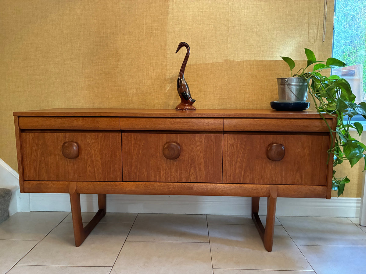 A mid-century modern wooden sideboard with three drawers and angled legs, styled in a home interior with a plant and a decorative sculpture on top.