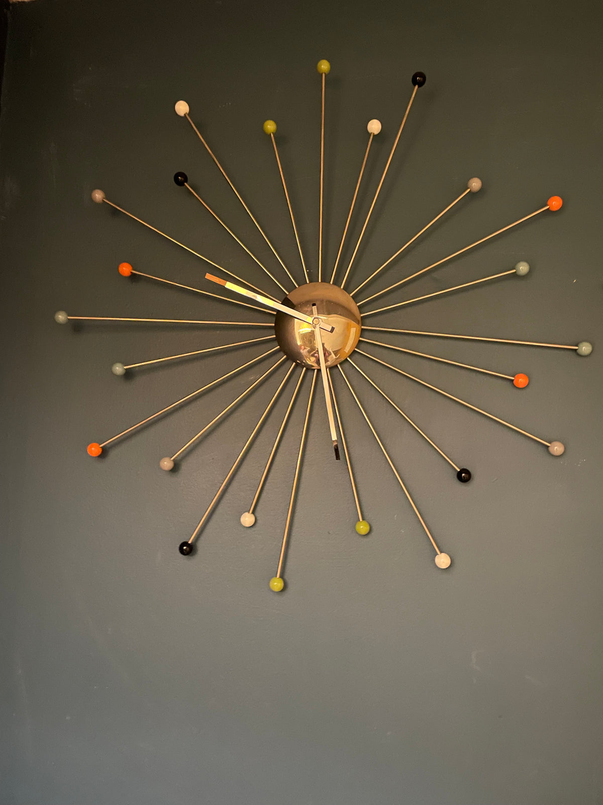 A mid-century modern starburst wall clock with a gold-tone metal center and multiple radiating rods ending in small colorful spherical tips, mounted on a dark gray wall.