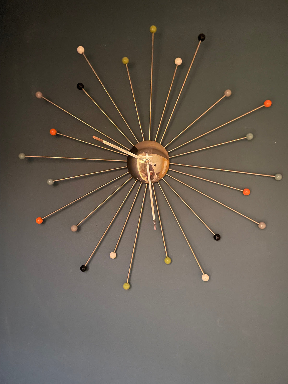 A mid-century modern starburst wall clock with a bronze-toned central hub and multiple thin metal rays ending in small colorful spherical tips, mounted on a dark gray wall.