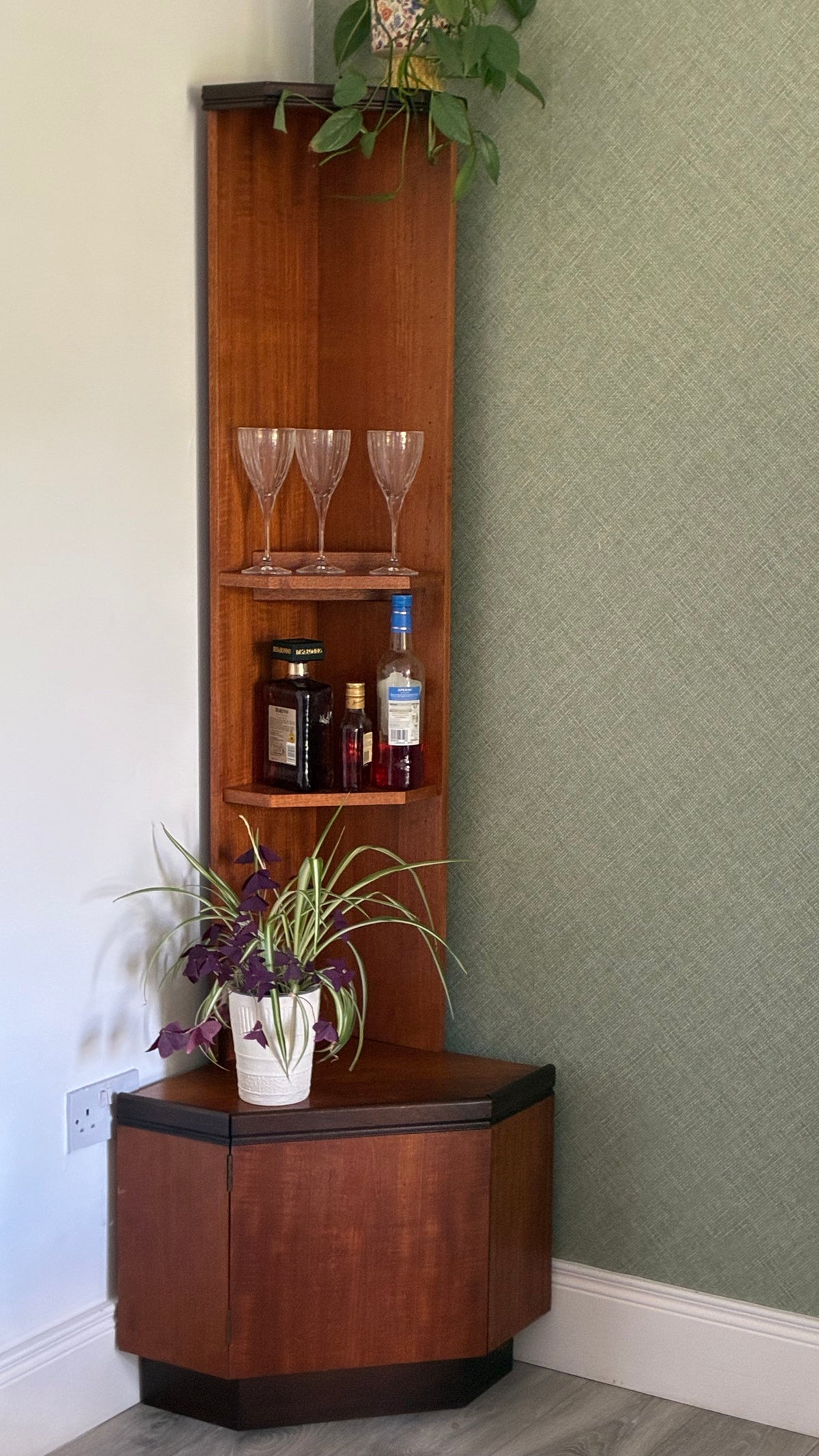 A corner wooden g plan cabinet with open shelving and a lower hexagonal storage base, styled with wine glasses, bottles, and potted plants in a room corner against a textured sage green wall.