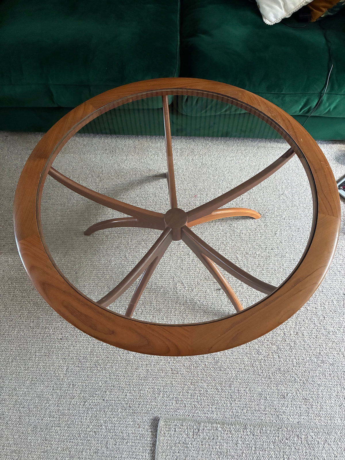 A mid-century modern g plan spider wooden coffee table with a clear glass top and curved star-shaped legs, positioned in a living room in front of a green velvet sofa.