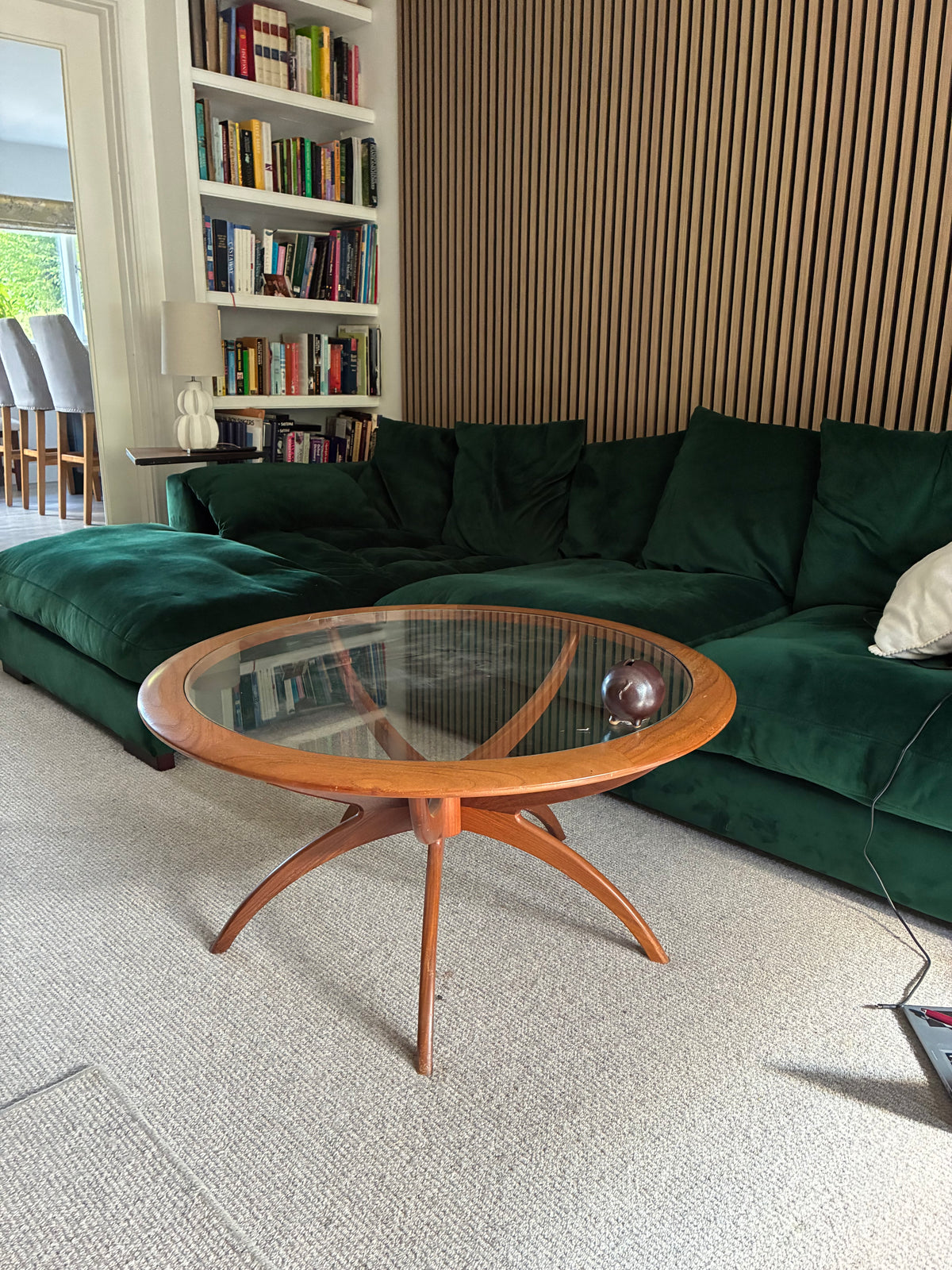 A mid-century modern g plan spider wooden coffee table with a clear glass top and curved star-shaped legs, positioned in a living room in front of a green velvet sofa.