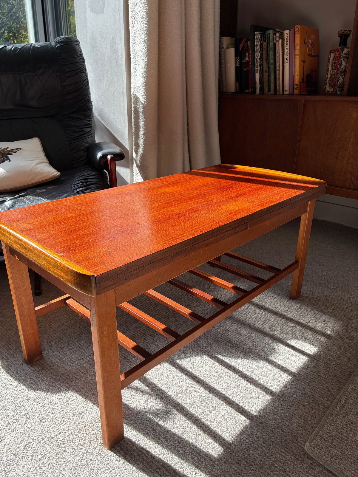 A rectangular wooden coffee table with a warm reddish-brown finish and a slatted lower shelf, photographed in a real living room setting with sunlight, a chair, curtains, and a bookcase in the background.