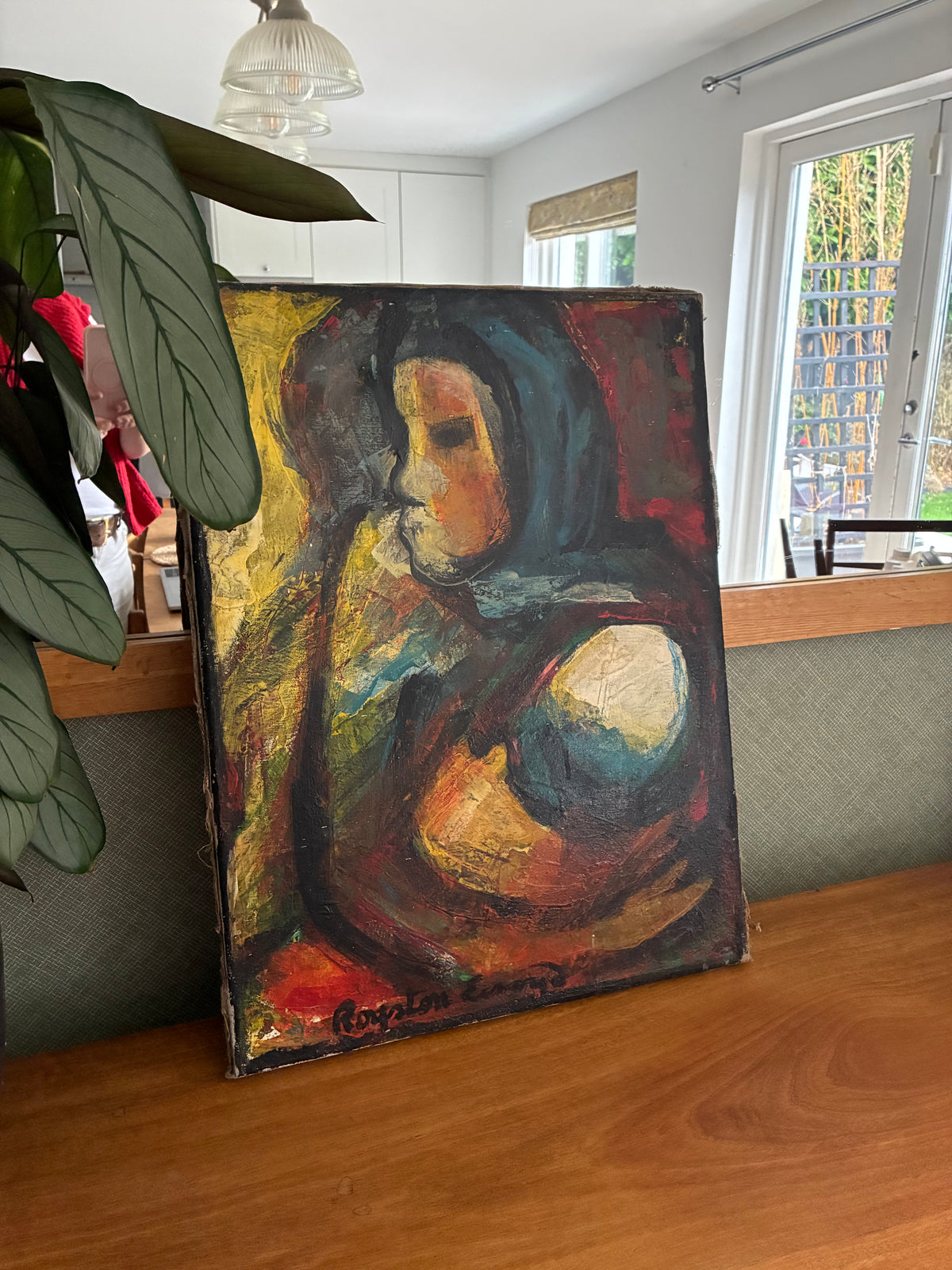 An expressive oil painting of a seated figure in profile, displayed on a wooden table in a home interior setting with a plant in the foreground.