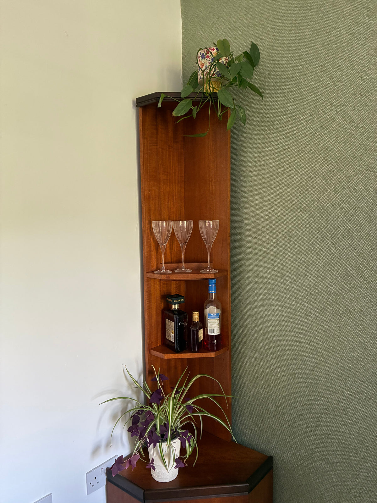 A tall, narrow wooden corner shelf with three tiers, styled with wine glasses, bottles, and potted plants in a home setting with a textured green wall and cream-colored wall.