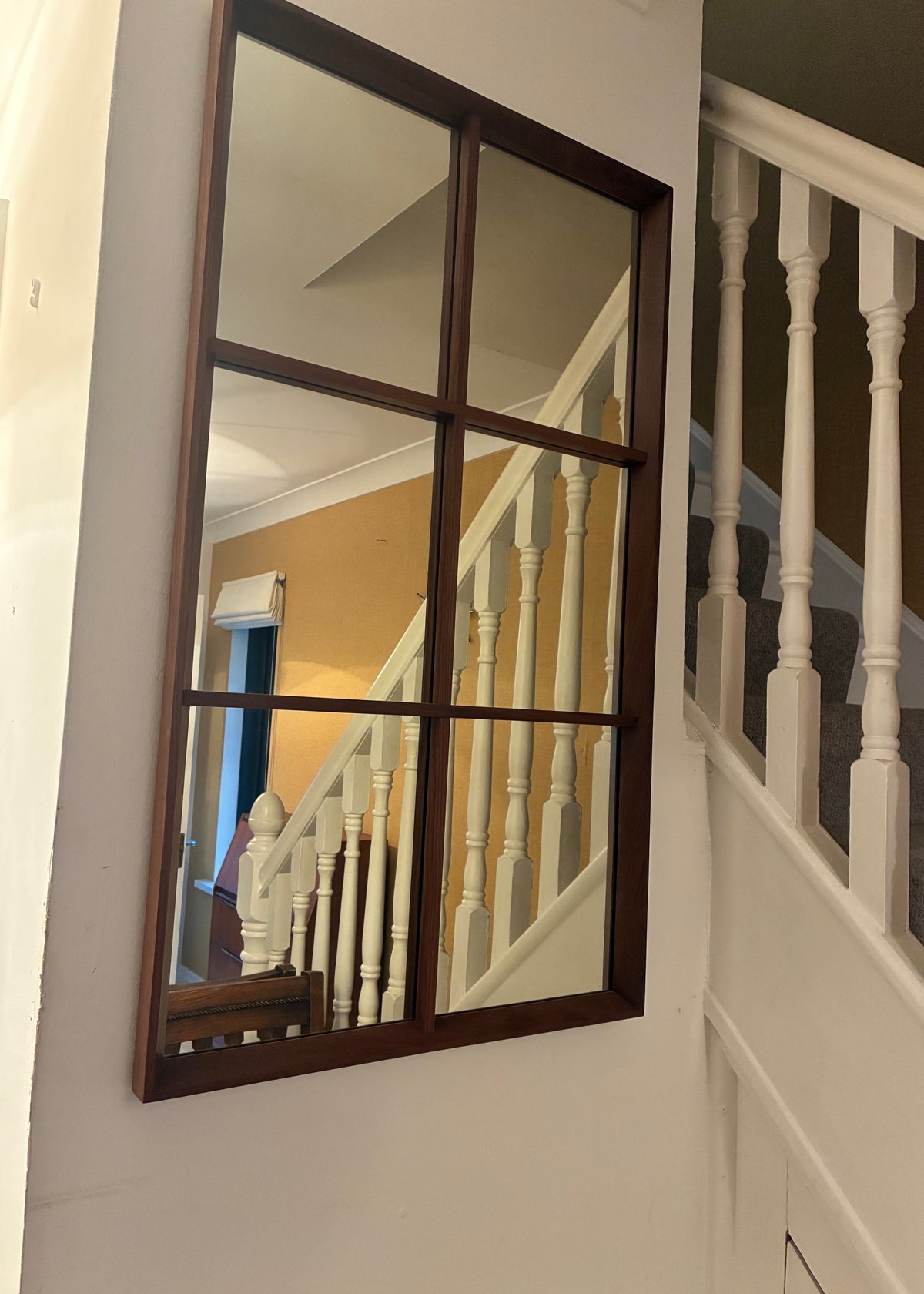 A large rectangular teak wall mirror with a dark wood frame hangs on a white wall beside a staircase with white balusters and a mustard-yellow wall.