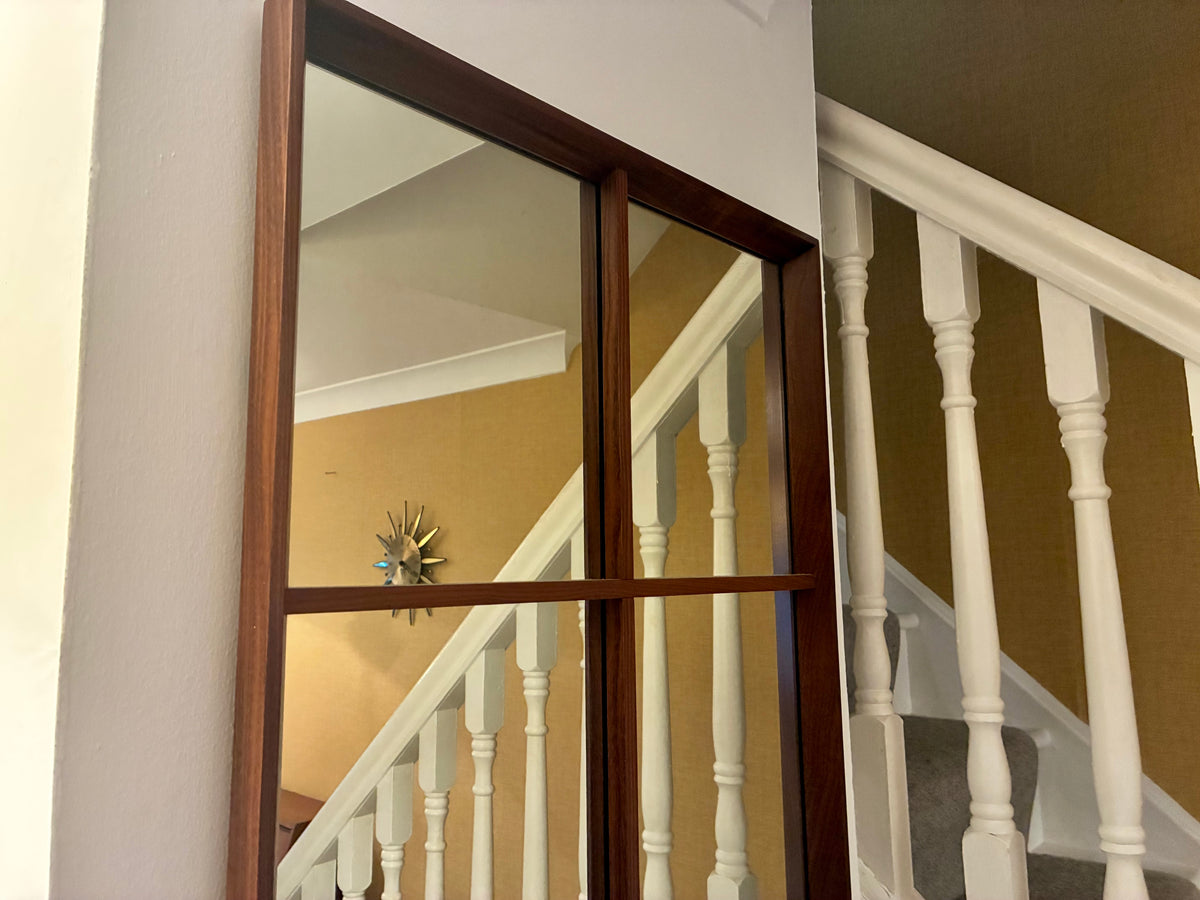 A large rectangular teak wall mirror with a dark wood frame hangs on a white wall beside a staircase with white balusters and a mustard-yellow wall.