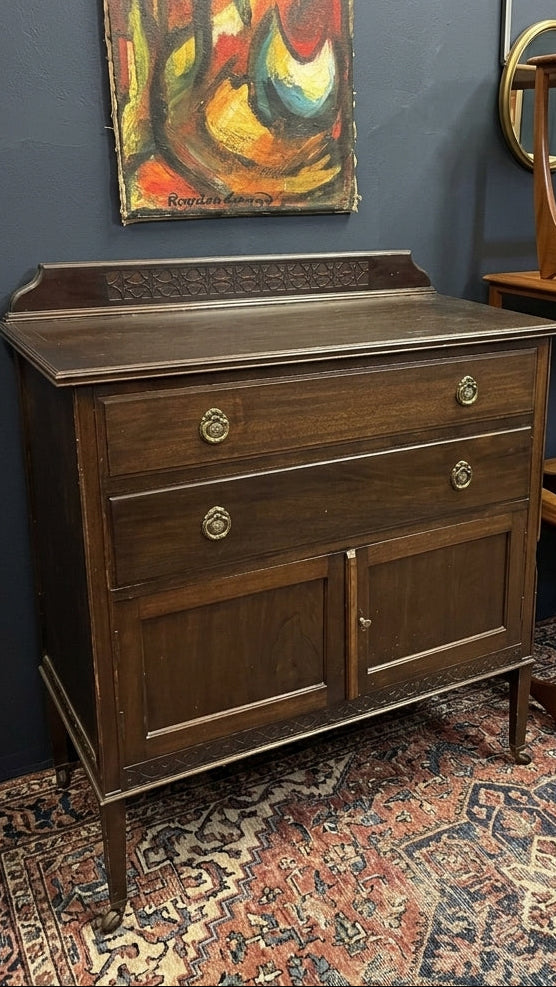 A dark brown wooden sideboard cabinet with two drawers and two lower doors, shown in a room setting against a dark blue-gray wall with an abstract painting and a patterned rug.