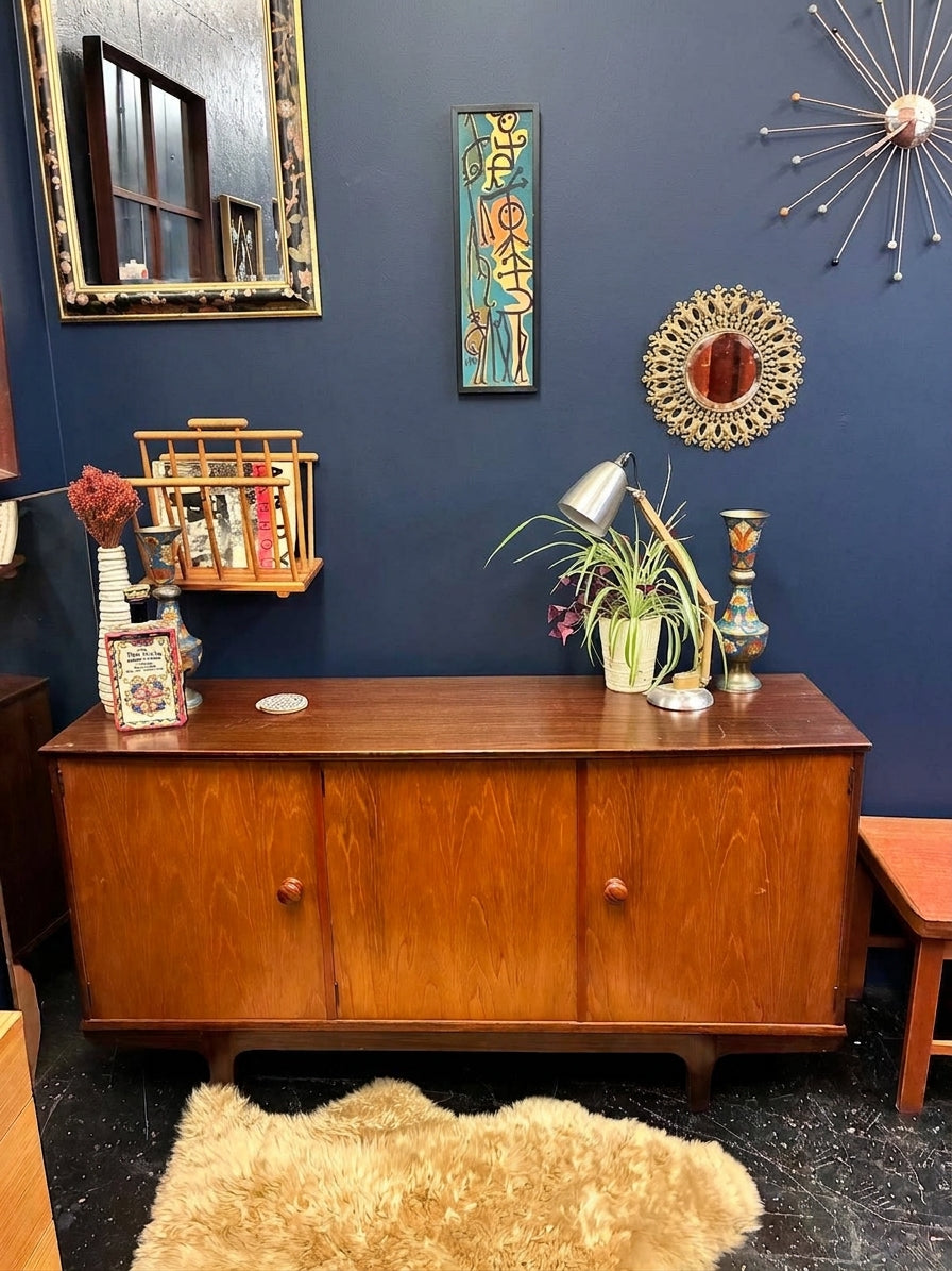 A mid-century modern wooden sideboard cabinet with three drawers, styled in a room with a dark blue wall, vintage mirrors, and home decor accessories.