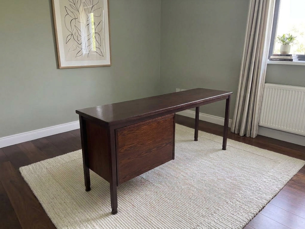 A dark brown wooden writing desk with a single drawer, positioned on a light beige woven area rug in a room with sage green walls and hardwood floors.