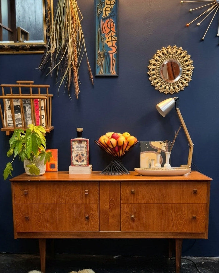 A mid-century modern wooden sideboard cabinet styled in a room with a dark blue wall, featuring decorative mirrors, a starburst wall light, a potted plant, and a bowl of fruit.